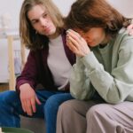 Crop male soothing disappointed crying female in sweatshirt and gray trousers on blurred background