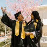Two students celebrating graduation with caps and gowns outdoors in spring.