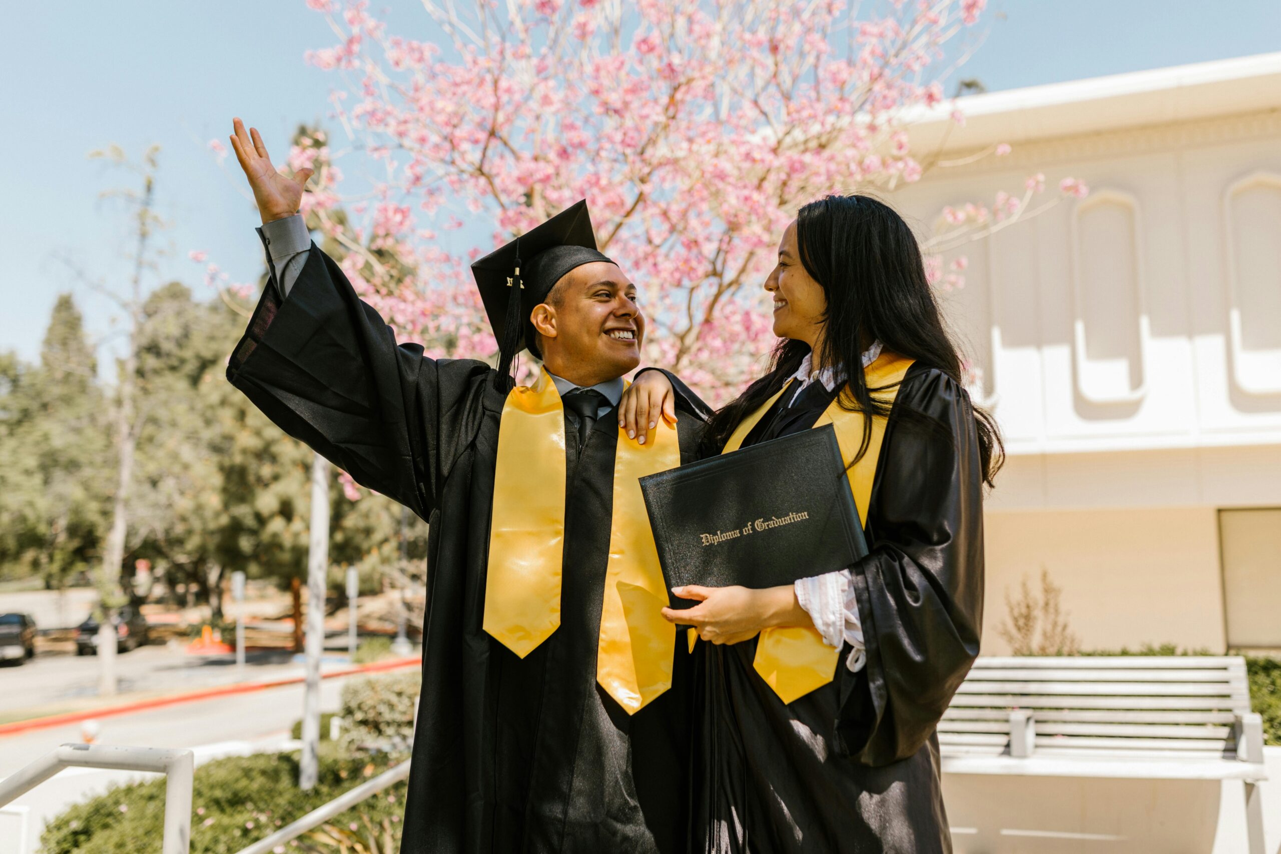 Two students celebrating graduation with caps and gowns outdoors in spring.